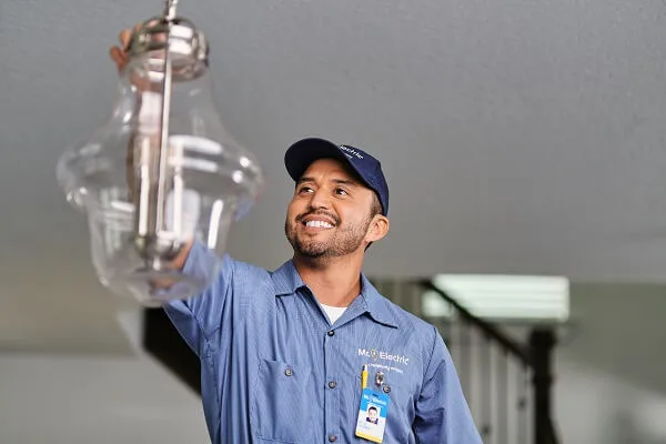 A smiling Mr. Electric service professional reaches for a pendant light fixture suspended from a ceiling